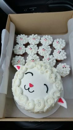 a cake in a box with white frosting and sheep faces on the top, surrounded by other cupcakes