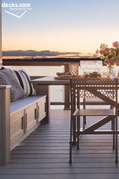 an outdoor table and chairs on a deck overlooking the water at sunset or dawn with flowers in vases