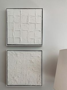 two white square paintings on a wall next to a lamp