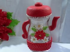 a glass teapot with red and white flowers on the lid, sitting next to a flower arrangement