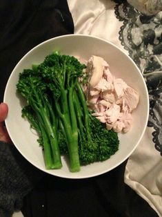 a person holding a white bowl with broccoli and chicken in it on a bed