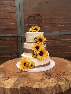 a three tiered cake with sunflowers on the top is sitting on a tree stump