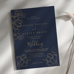 a blue and gold wedding card with flowers on the front is laying next to a white flower