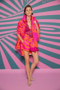 Vivienne Rohner stands against a pink and turquoise swirly background modeling Brandon Maxwell's Spring Summer 2022 Look 3. The outfit consists of board shorts and a hooded parka in the same orange and fuchsia swirl pattern, along with two multicolored beaded choker necklaces with a centered silver butterfly pendant and metallic green pumps. The model has a short brown bob with one hand on the strap of an orange and fuchsia backpack that is hanging on her left shoulder. Vivienne Rohner, New Year Look, Tangerine Dress, Brandon Maxwell, Cocktail Attire, Spring Summer 2022, Knitwear Fashion, Blair Waldorf, Hooded Parka