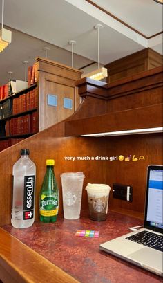 a laptop computer sitting on top of a wooden counter next to cups and drinks in front of bookshelves