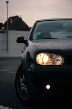 the front end of a black car with its headlights turned on and it is parked in a parking lot