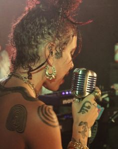 a woman with tattoos on her arm holding a microphone and looking at the camera while standing in front of a crowd