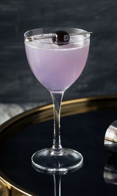 a glass filled with purple liquid and topped with a black olive on a gold tray