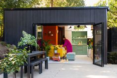 a woman sitting at a table in front of a black shed with plants and potted plants