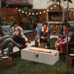 group of people sitting around a fire pit in the grass with lights on it and one person lighting something