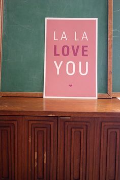 a pink poster with the words la la love you in front of a green chalkboard