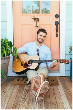 a man sitting in front of a pink door holding a guitar and smiling at the camera