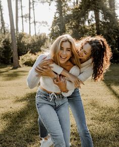 two young women hugging each other in the grass with trees in the back ground behind them