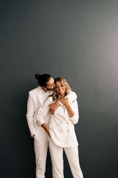 a man and woman in white suits posing for a photo against a gray wall with their arms around each other