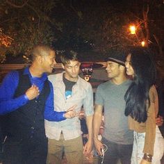 four people standing around talking to each other in the street at night with trees and lights behind them