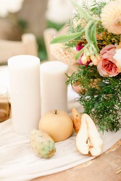 an arrangement of flowers, candles and fruit on a white table cloth with a vase in the background