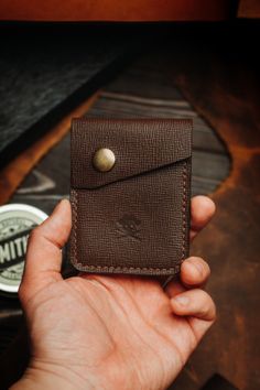 a hand holding a brown leather wallet on top of a wooden table next to a cup