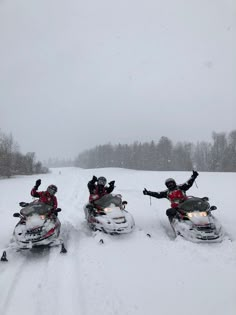 three people riding snowmobiles on a snowy road with trees in the background and one person waving