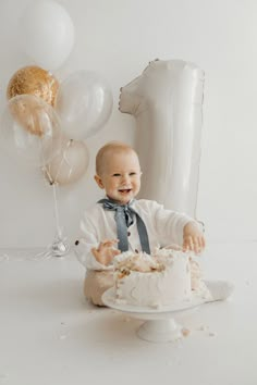 a baby sitting in front of a cake with balloons behind it and a white background