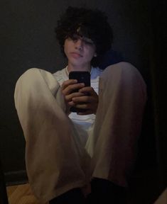 a young man sitting on the floor looking at his cell phone while wearing white shirt and black pants