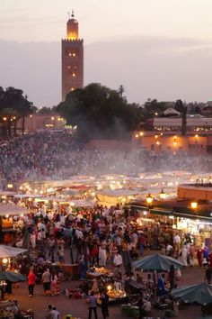 an outdoor market with lots of people standing around it at night and in front of a clock tower