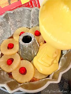 someone is pouring yellow liquid on pineapple slices in a metal bowl with red cherries