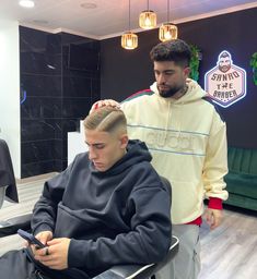 a man sitting in a barber chair while another man looks at his cell phone as he gets his hair cut