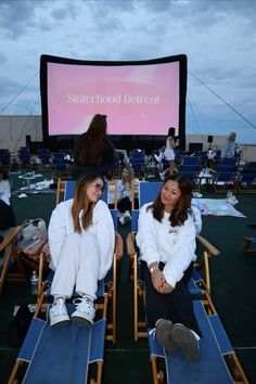 two women are sitting in lawn chairs watching a movie on the big screen at sunset