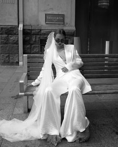 a woman sitting on top of a wooden bench in front of a building wearing a wedding dress