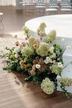 flowers and greenery are arranged on the floor next to a white round table with chairs in the background