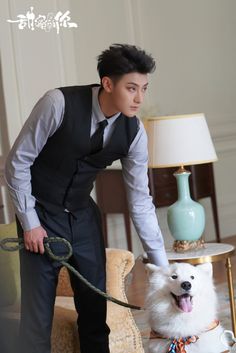 a man in a black vest and tie holding a leash to a white dog on a couch