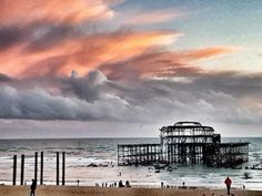 people are walking on the beach as the sun sets in front of an old pier