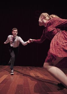 a man and woman dance on a wooden floor in front of a dark background, with one holding the other's hand