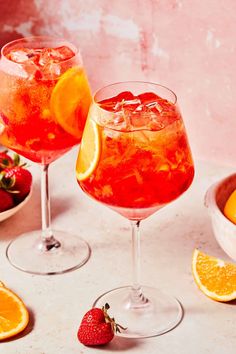 two glasses filled with fruit and ice next to bowls of strawberries, oranges and strawberries