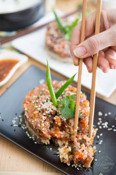 a person holding chopsticks over some food on a black plate with rice and sauce