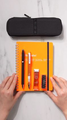 a person holding an orange notebook with pens and pencils in it next to a black case