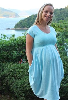 a pregnant woman in a blue dress standing by bushes and looking at the camera with her hands on her hips