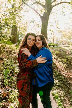 two women hugging each other while standing in front of a tree with leaves on it