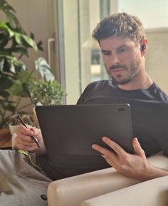 a man sitting on a couch holding a tablet computer and writing something in his hand