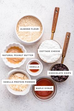 ingredients to make chocolate chip cookies laid out on a white counter top, labeled in the words almond flour, cottage cheese, butter, vanilla protein powder, and maple syrup