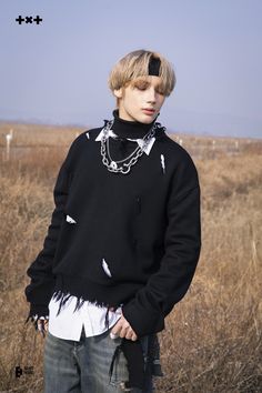 a young man standing in a field wearing a black sweater with torn edges and a chain around his neck