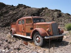 an old rusted out truck is parked in the dirt near some rocks and trees