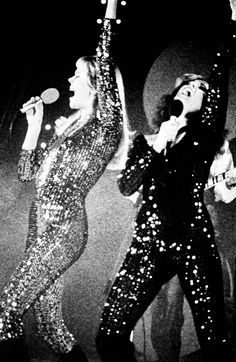 two women in sequin jumpsuits on stage with one holding her arms up