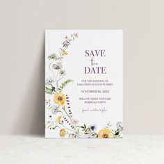 a floral save the date card on top of a white table with purple and yellow flowers