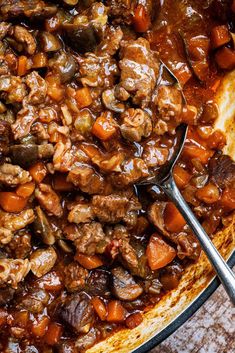 a large pot filled with meat and carrots on top of a wooden table next to a spoon