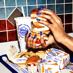 two hands holding hamburgers and french fries in front of a tray of food on a table