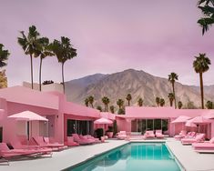 a pool with lounge chairs and umbrellas next to palm trees in front of a pink building