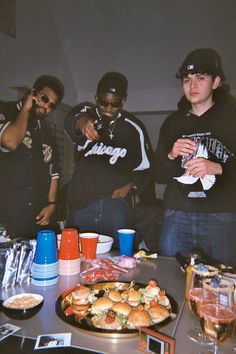 three young men standing around a table with food on it and drinks in front of them