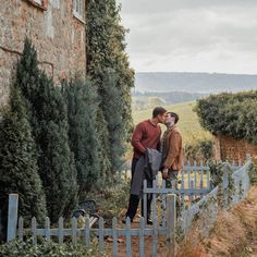 two people standing next to each other in front of a stone building with trees and bushes