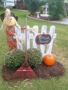 a scarecrow holding a broom in front of a white picket fence and pumpkins